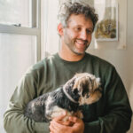 Smiling man in a green sweater holding a small fluffy dog indoors by a window.